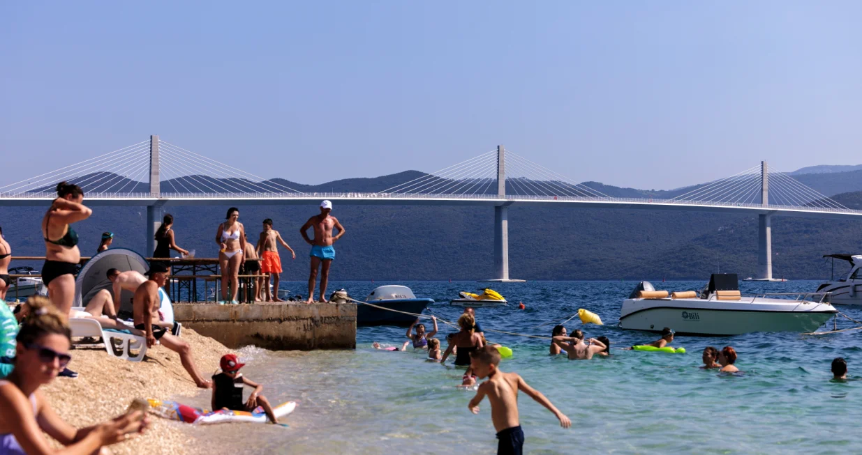 epa10092340 People enjoy at the beach, with the Peljesac Bridge in the background, at Komarna village, Croatia, 26 July 2022. Two and a half years ago started the construction of a new bridge to achieve the territorial continuity of the country by connecting the southern exclave compromising the bulk of Dubrovnik-Neretva County with the remainder of the Croatian mainland and to avoid using the Bosnian territory to cross from one part of Croatia to another. Croatian authorities are opening the bridge on 26 july. EPA/STRINGER/Stringer