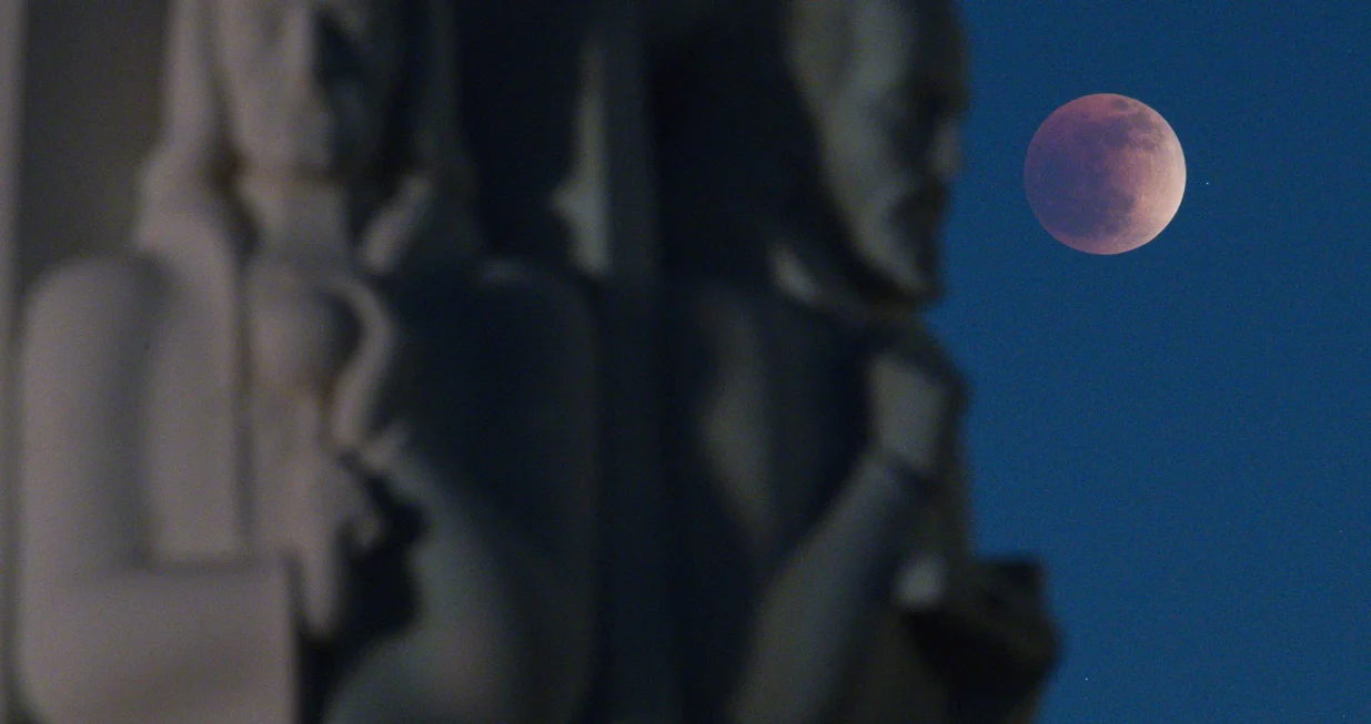 epa09950171 The Super Flower Blood Moon lunar eclipse peers behind the 'Astronomers Monument', including statues of Johanees Kepler (R) and Isaac Newton (L), at the Griffith Observatory in Los Angeles, California, USA, 15 May 2022. EPA/CAROLINE BREHMAN/Caroline Brehman