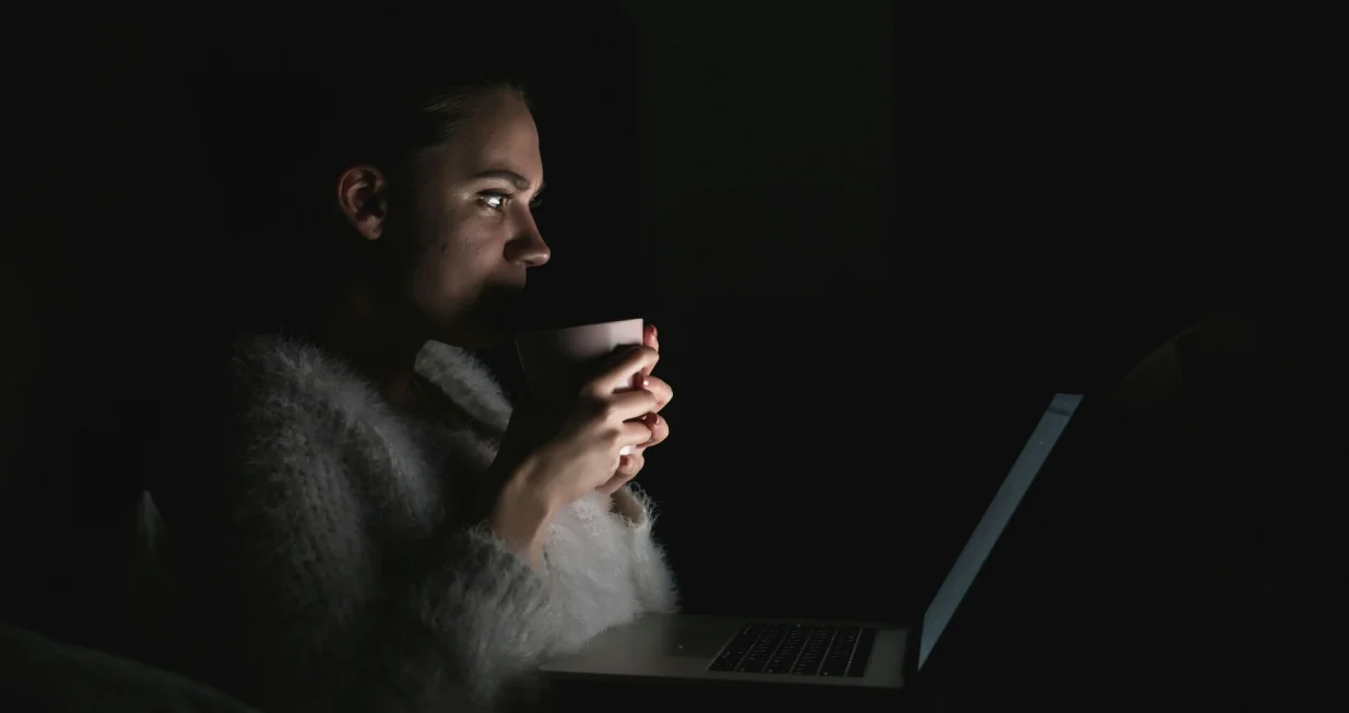 Djevojka leži u krevetu noću i pažljivo gleda u ekran laptopa. Jede hranu, drži šolju i gleda seriju noću./Anastasiia Nelen