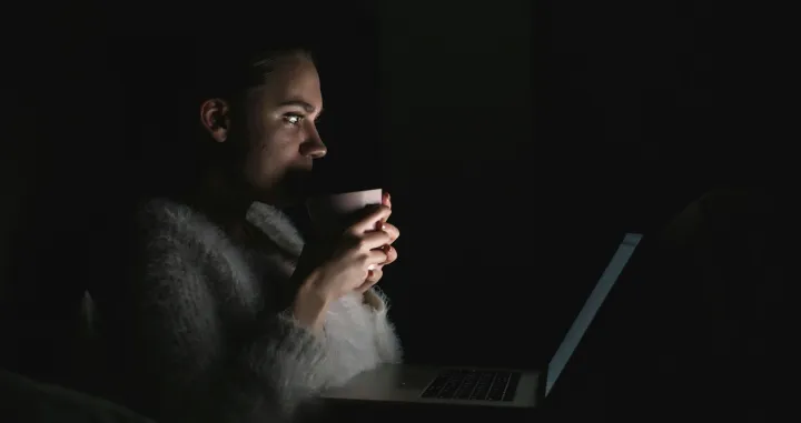 Djevojka leži u krevetu noću i pažljivo gleda u ekran laptopa. Jede hranu, drži šolju i gleda seriju noću./Anastasiia Nelen