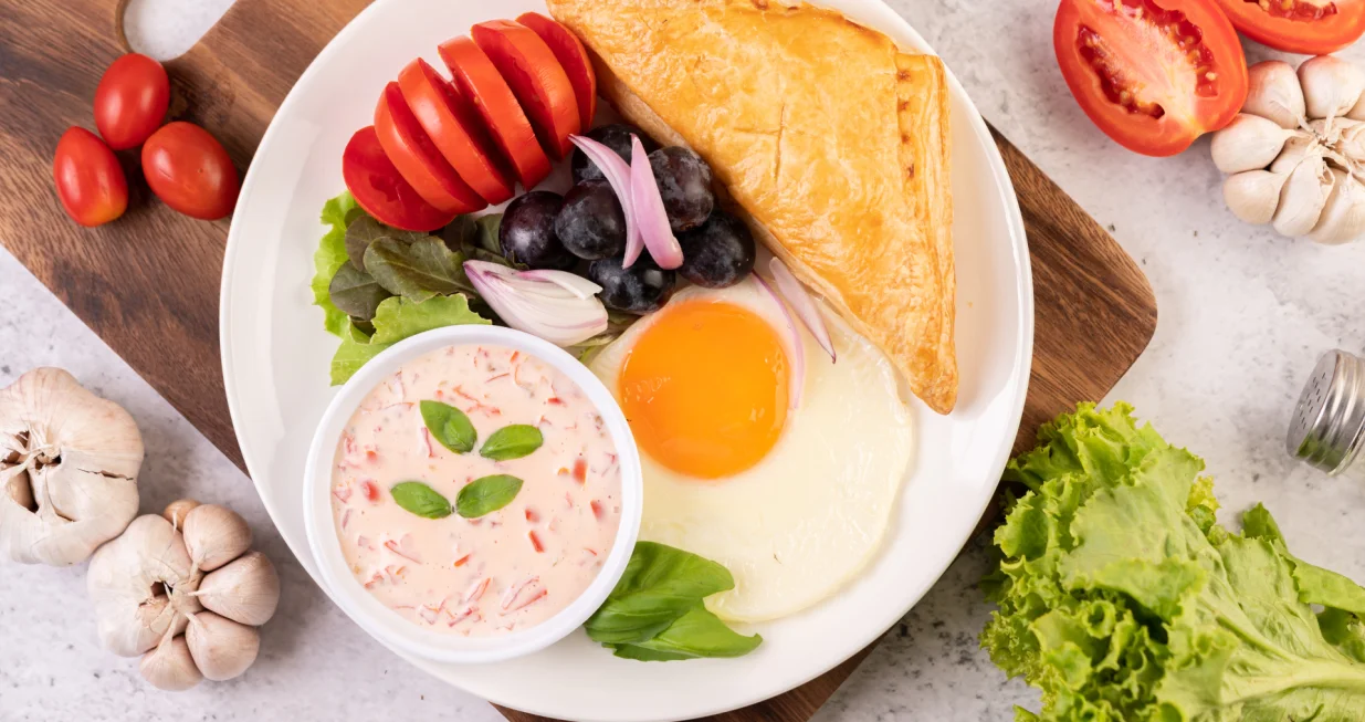 Breakfast consists of bread, fried egg, salad dressing, black grapes, tomatoes, and sliced ​​onions./Freepik