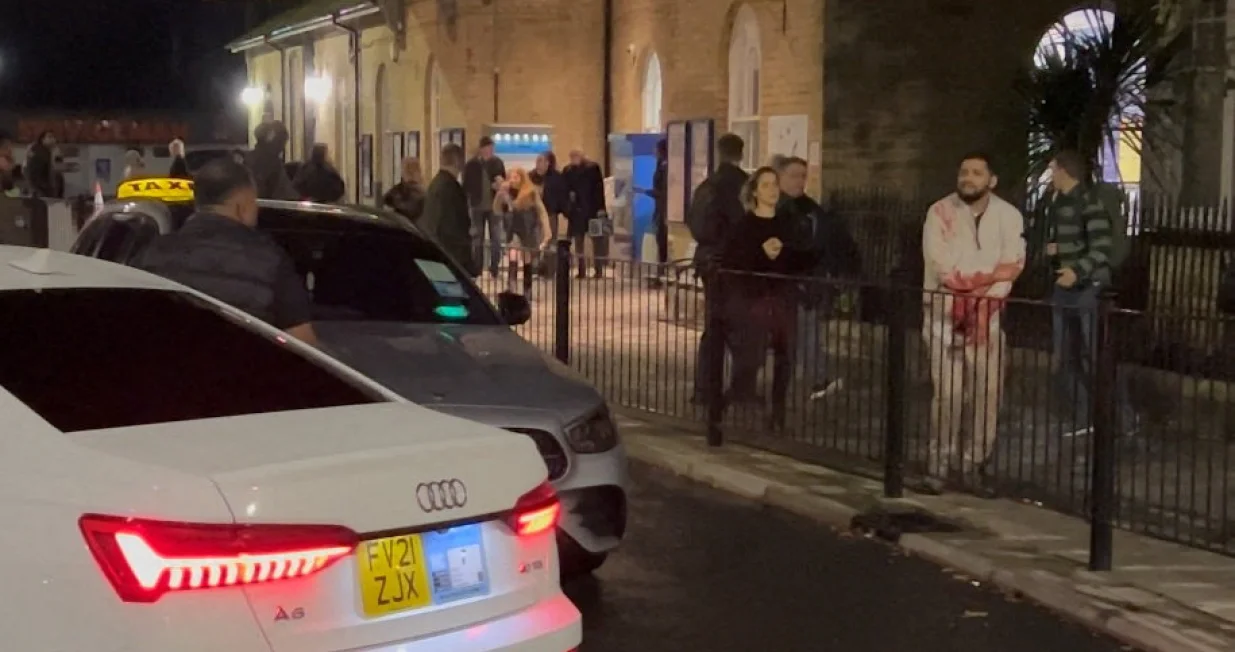 A wounded man walks on a sidewalk following a stabbing incident at a train station in Huntingdon, Britain, November 1, 2025, in this screengrab obtained from a social media video. @_ACC5_/via REUTERS THIS IMAGE HAS BEEN SUPPLIED BY A THIRD PARTY. MANDATORY CREDIT. NO RESALES. NO ARCHIVES. THIS IMAGE WAS PROCESSED BY REUTERS TO ENHANCE QUALITY. AN UNPROCESSED VERSION HAS BEEN PROVIDED SEPARATELY. VERIFICATION: Reuters was able to confirm the location and date of the footage by original file metadata from the source./@_acc5_