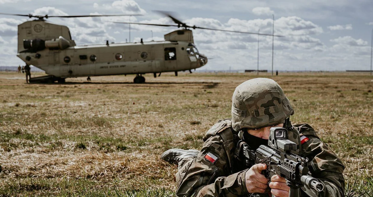 A Polish Armed Forces Soldier scans his sector as a U.S. Army CH-47 Chinook helicopter from the 1-214th General Support Aviation Battalion, 12th Combat Aviation Brigade, loads up personnel in the background, during a joint exercise led by the Polish Armed Forces at the landing zone near the G2A Arena, Poland, April 8, 2022. This exercise and others like it enhances our interoperability with our NATO allies and partners and strengthens the regional relationships that we have developed. 12 CAB is the only enduring aviation brigade present throughout Europe that enables us to deter and defend against threats from any direction. 12 CAB is among other units assigned to V Corps, America's Forward Deployed Corps in Europe that works alongside NATO Allies and regional security partners to provide combat-ready forces, execute joint and multinational training exercises, and retains command and control for all rotational and assigned units in the European Theater.(U.S. Army photo by Staff Sgt. Thomas Mort)/Staff Sgt. Thomas Mort