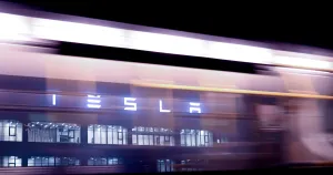 epa10387461 The Tesla logo seen through a window of a bus passing in front of the Tesla Gigafactory in Gruenheide near Berlin, Germany, 03 January 2023. The Tesla Gigafactory Berlin-Brandenburg plant in Gruenheide produces 2,000 Model Y electric cars in a week. EPA/Filip Singer/Filip Singer